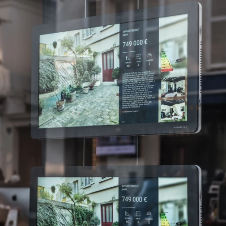 Zdjęcie przedstawia nowoczesny system linkowy LCD, wykorzystywany w witrynie biura nieruchomości. Na ekranie widoczna jest prezentacja cyfrowa z ofertą sprzedaży mieszkania, zawierająca zdjęcia nieruchomości, cenę oraz kluczowe informacje techniczne. System charakteryzuje się eleganckim designem, podkreślając profesjonalizm ekspozycji.